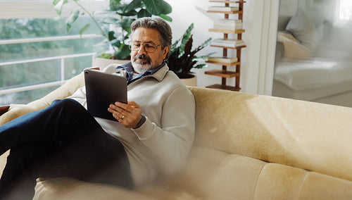 Senior man uses tablet while relaxing on sofa at home