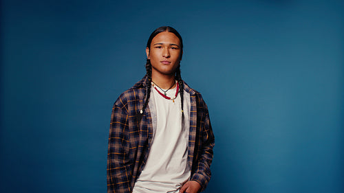 Young man laughs in studio against blue backdrop with braided hair and plaid shirt
