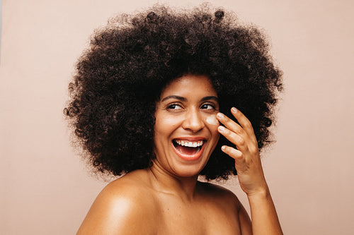 Excited woman with Afro hair applying cosmetic moisturizer on her face