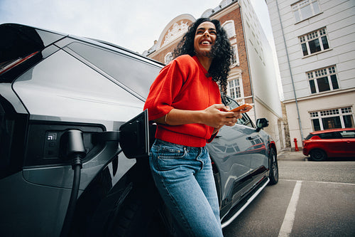 Joyful woman uses smartphone while charging electric car in city