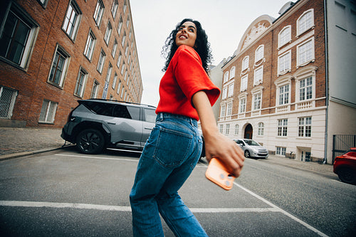 Woman walking to electric car charger with phone on urban street