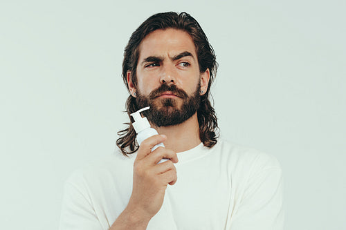 Man holding facial moisturizer with a thoughtful expression in a studio