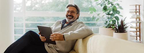 Senior man smiles while using a tablet on the sofa