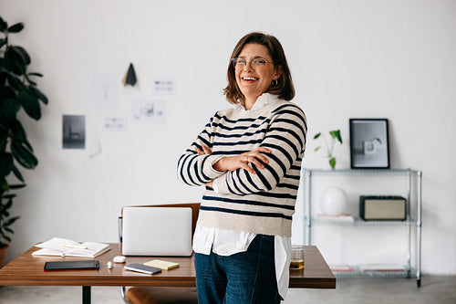 In the workspace, the woman stands confidently by her desk with a laptop for business leadership
