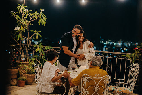 Man embracing wife while parents look on proudly during evening balcony gathering