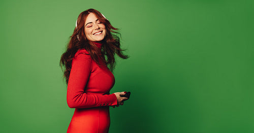Fashionable woman with red hair, smiling and holding smartphone with headphones on a green studio background