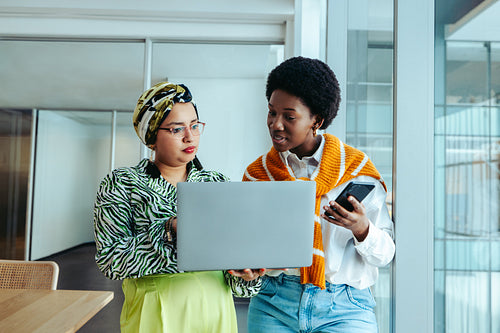 Female coworkers discussing a design brief in office with a laptop and smartphone