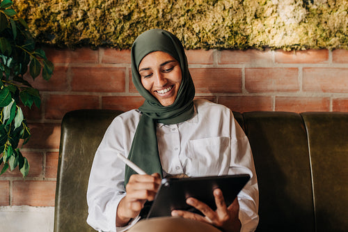 Woman with a hijab smiling happily while drawing on a digital tablet