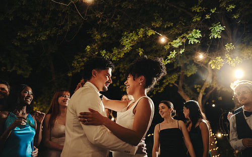 Romantic outdoor wedding dance under string lights surrounded by friends