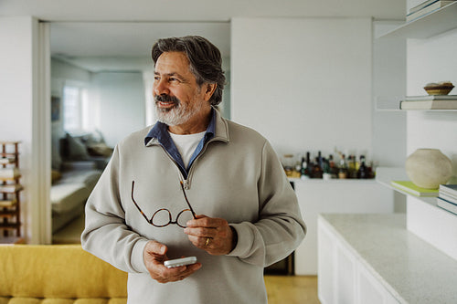 Grey-haired senior holds a smartphone and glasses at home