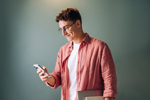 Young man using smartphone while holding a laptop and smiling