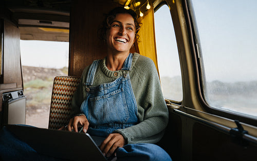 Woman smiles while working on a laptop inside a cozy camper van on a road trip