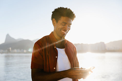 Cheerful man texting on smartphone at sunset by the water