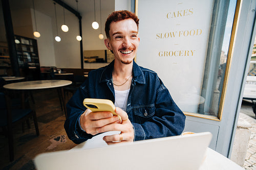 Smiling man in cafe using smartphone and laptop