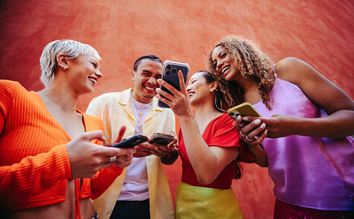 Laughing friends browse smartphone content together outdoors against an orange wall