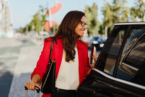 A businesswoman entering car with suitcase and phone during a bright sunny day