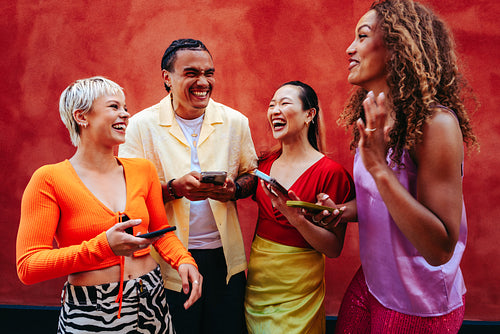 Chatting friends share smartphone content and laugh together against a bright red wall