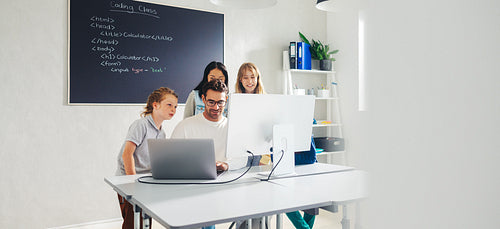 Elementary school teacher leading a coding lesson in a classroom, using a computer to show his students how to code