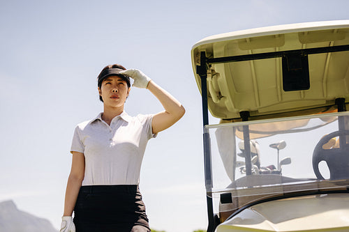 Confident woman golfer shielding her eyes from the sun, standing next to a golf cart