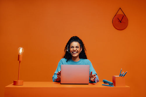 Young female sales representative laughing while working on a laptop against a vibrant orange background