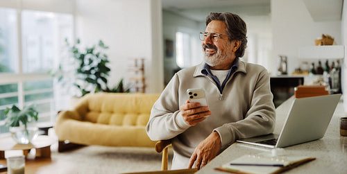 Senior man uses smartphone with laptop at home
