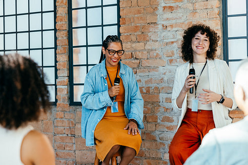 Women giving a speech at a seminar with a brick wall background, smiling and holding microphones