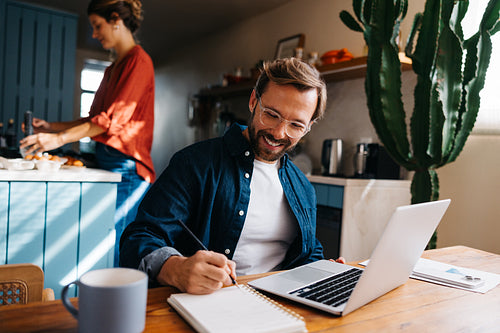 Man at home working on laptop and writing in notebook