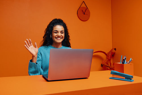 Female remote worker waving while working on laptop in vibrant orange office