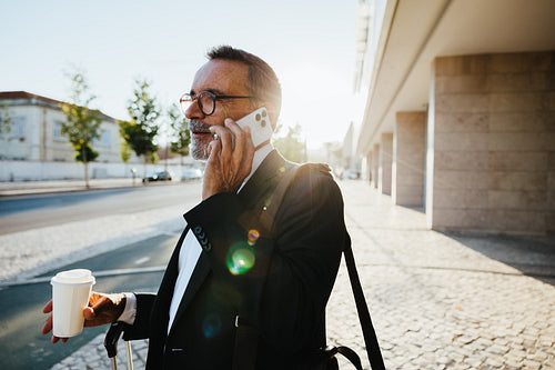 Senior man in suit using phone with coffee outdoors in urban setting