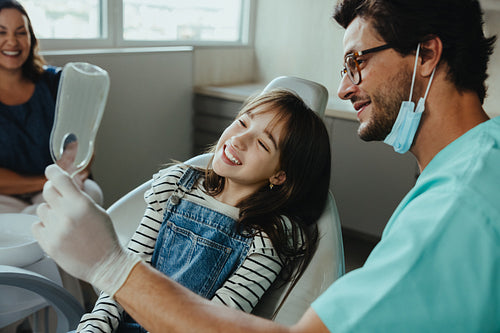Dentist uses mirror to show young patient her teeth after checkup