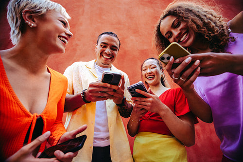 Friends laugh over smartphone content together outdoors by a bright wall