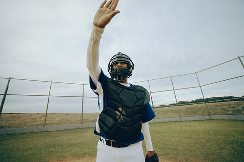 Baseball catcher wearing chest protector and shin guards, signaling with hand raised for teamwork and focus