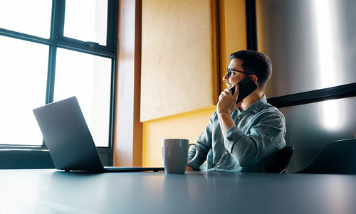 Young professional man talking on phone while working on laptop in modern office