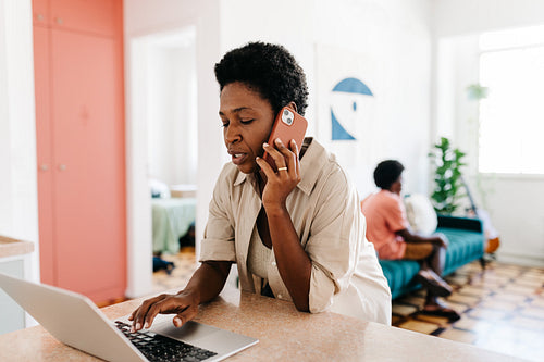 Working mom speaking on the phone while using a laptop in a home kitchen