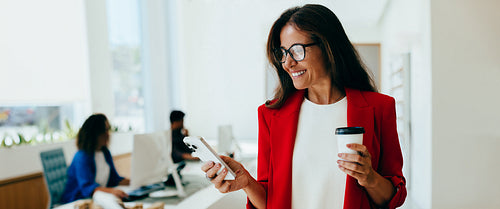 Confident businesswoman checking smartphone with coffee in hand at the office