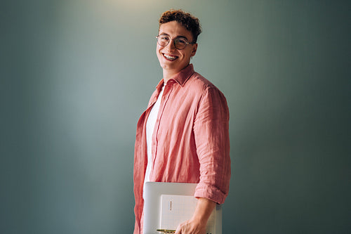 Smiling young man in casual attire holding a notebook against a teal background