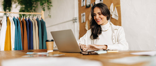 Fashion designer happily working in her boutique, using a laptop to create her clothing designs