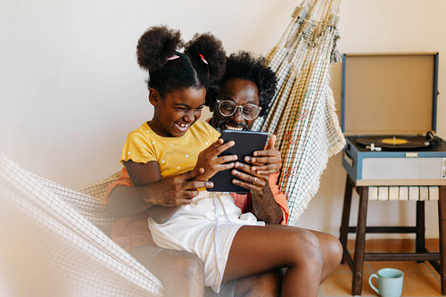 Dad and daughter relaxing together in hammock, laughing and watching a video on a tablet