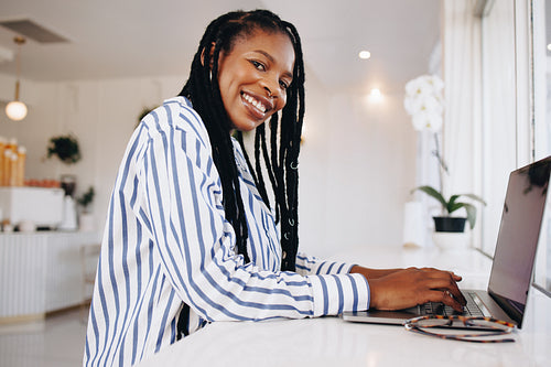 Cheerful female freelancer smiling at the camera while typing on a laptop in a cafe