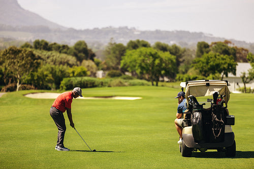 Man aiming for the green near sand bunker, with friend relaxing in golf cart