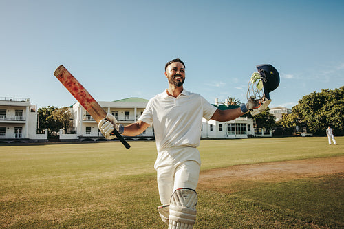 Cricketer celebrating achievement holding bat and helmet on a sunny field