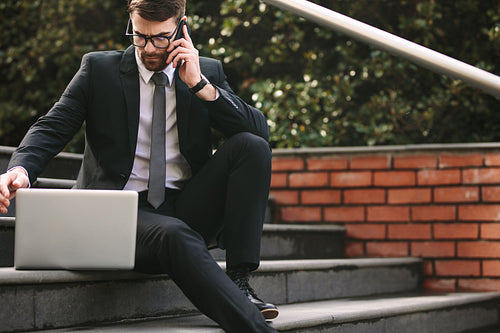 Businessman sitting on steps with laptop talking over phone
