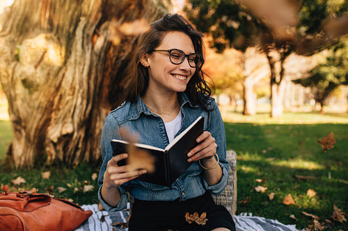 Smiling woman sitting at park with book and leaves falling around