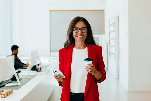 Confident woman in red jacket holding coffee in bright office environment