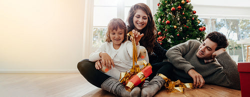 Family opening gifts by the Christmas tree on Christmas morning - kid excited about unwrapping a present from her parents.