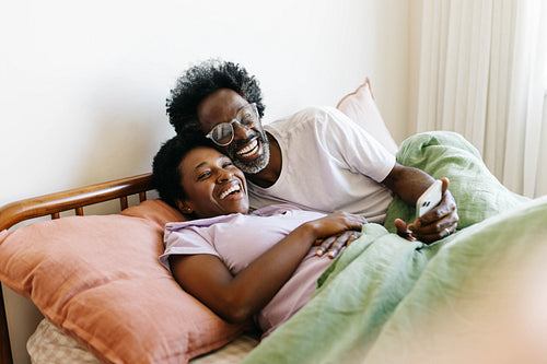 Quality time in a relaxed home: Couple laughing and browsing social media together in bed