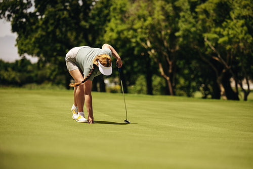 Woman golfer crouching on putting green with golf club preparing a shot in sunny weather