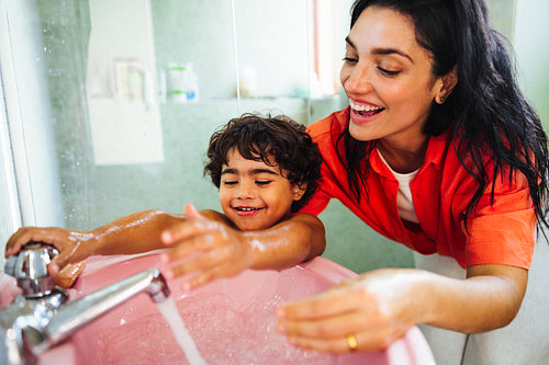 Mother helping joyful child wash hands at a sink with running water