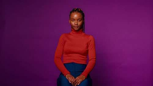 Young woman with braids sits calmly on a stool against a vibrant purple backdrop