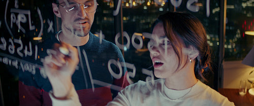 Focused business partners work late, solving an equation on a window overlooking the city